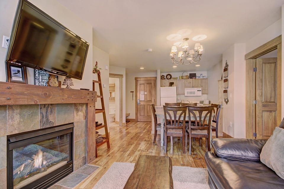 A cozy living room with a fireplace, wall-mounted TV, and wooden furniture. The open-plan space leads to a connected dining area with six chairs and a kitchen equipped with white appliances.