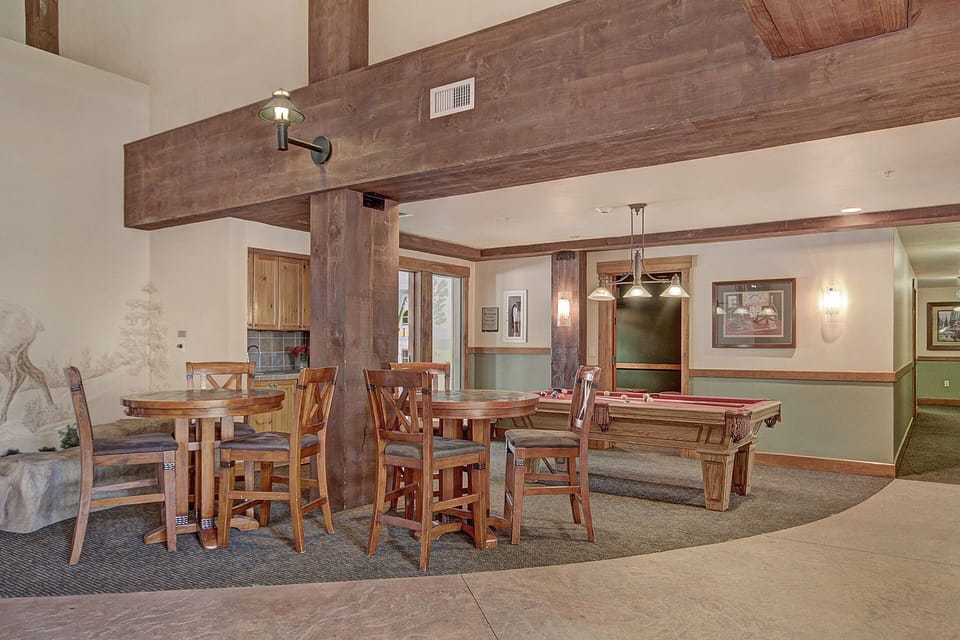 A rustic recreation room with wooden tables and chairs, a pool table under pendant lights, and artwork on the walls.