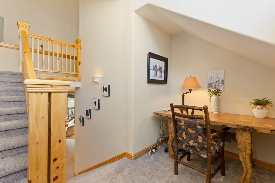 A small workspace with a wooden desk and chair situated on a carpeted landing. Stairs with wooden railings are to the left. The desk is decorated with a lamp, a plant, and a framed wall picture.