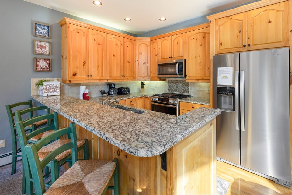 A kitchen with wooden cabinets, stainless steel appliances, a granite countertop, and a breakfast bar with green chairs. The space is well-lit and the refrigerator has a paper note attached to it.