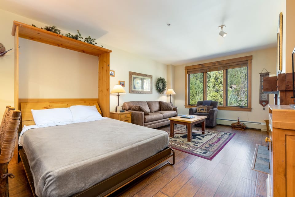 A cozy living area with a drop-down bed, couch, coffee table, and a large window showcasing a forest view. The room has wooden floors, rustic decorations, and a small fireplace.