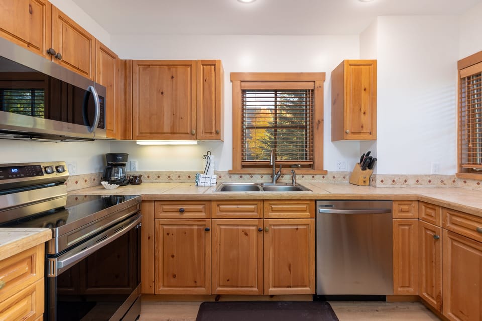 A modern kitchen with wooden cabinets, a stainless steel stove, microwave, and dishwasher, a double sink, and a window with wooden blinds.