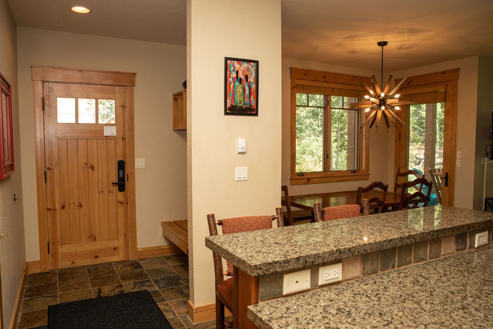 A cozy home interior with a granite kitchen countertop in the foreground, a wooden dining table with chairs, and a wooden entry door with a small window pane in the background.