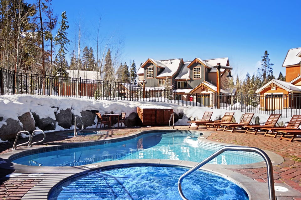 A view of an outdoor swimming pool and hot tub surrounded by snow with lounge chairs on the pool deck and houses in the background under a clear blue sky.