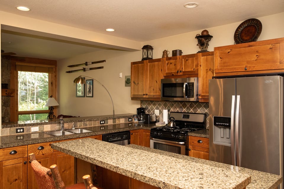 A modern kitchen features wooden cabinets, granite countertops, stainless steel appliances, and a double sink. An open window reveals a view of greenery outside. Cozy lighting adds warmth to the space.