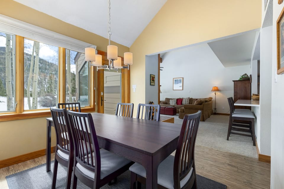 A dining room with a dark wooden table and six chairs, adjacent to a living area with a brown sofa and a wooden desk. A large window and a light fixture are visible.