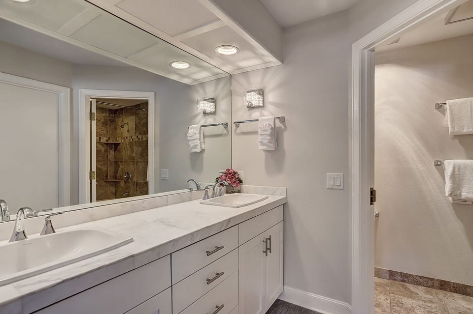 Modern bathroom with a marble countertop double sink vanity, wall-mounted towel racks, and a spacious shower area with a glass door. The lighting is bright with fixtures above the mirror.