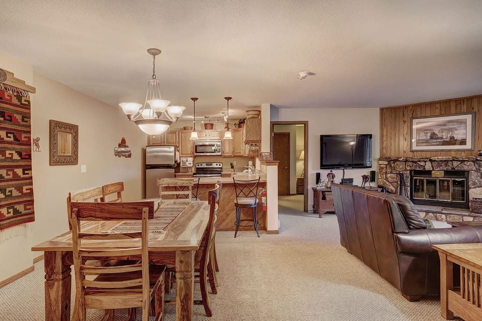 A cozy living area with a dining table, kitchen with stainless steel appliances, and a seating area featuring a leather couch, flat-screen TV, and stone fireplace. The room is lit by a chandelier.