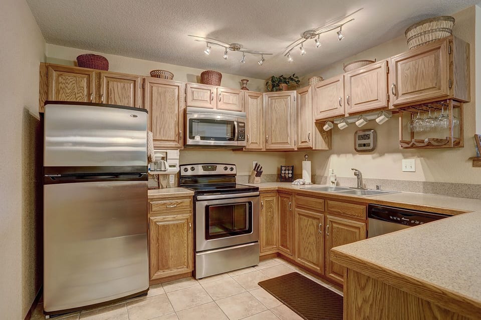 A kitchen with stainless steel appliances, wooden cabinets, tiled flooring, and a beige countertop. There are various kitchen items like a coffee maker, baskets, and utensils placed on the counters and shelves.