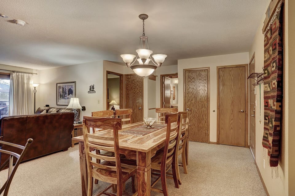 A dining room with a wooden table set for six, under a modern chandelier. Adjacent is a living area with a brown sofa. A hallway with closed wooden doors is visible in the background.