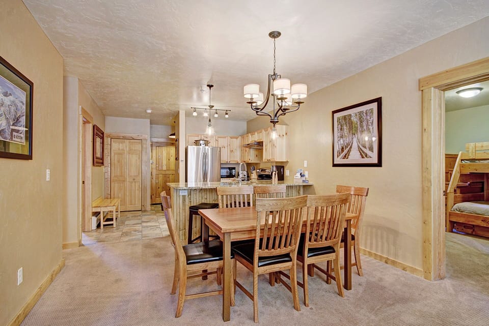 A cozy dining area with a wooden table and six chairs is adjacent to a bright kitchen. The room features beige walls, framed artwork, and modern light fixtures. A bunk bed is visible in the background.