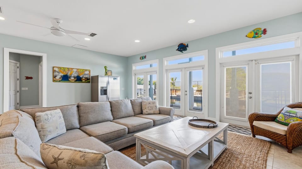 Light blue walls frame a sectional sofa and wicker chair facing a white wooden coffee table near glass doors with ocean views.