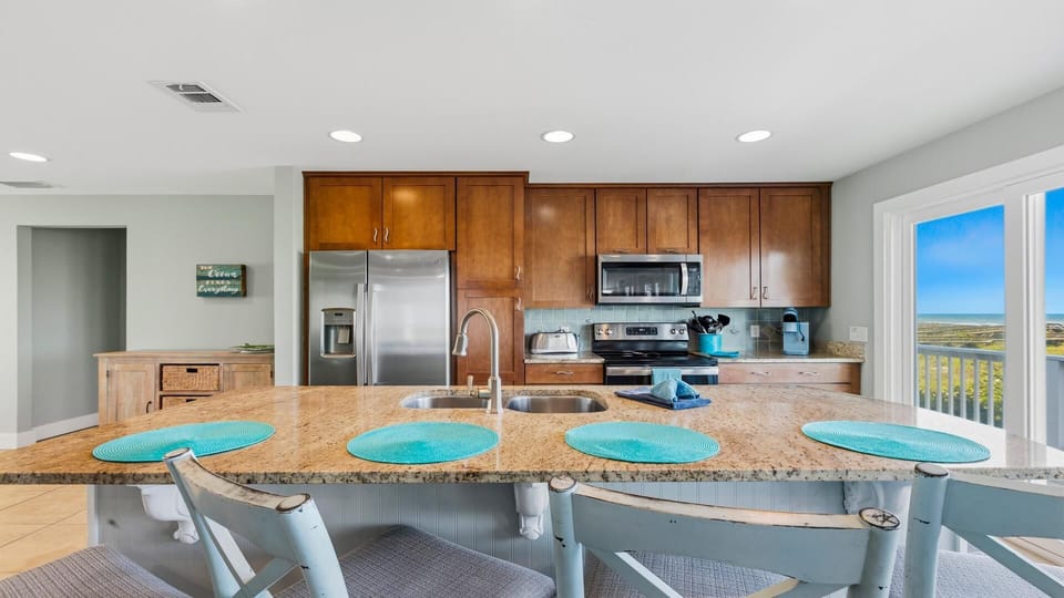 Granite kitchen island with double sink and distressed blue wooden stools faces sliding doors with ocean views.