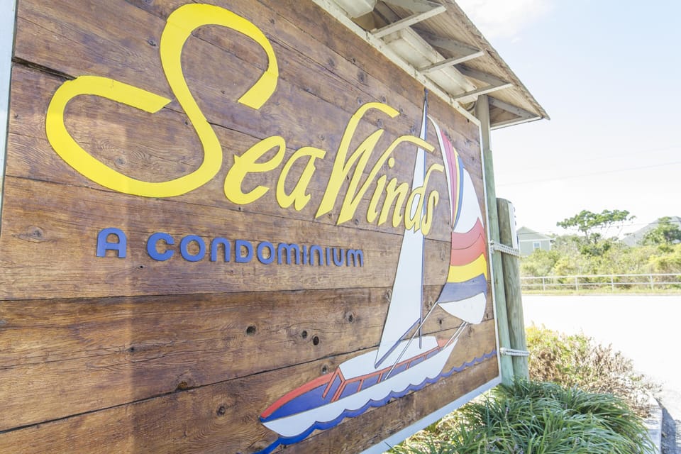 Wooden entrance sign with yellow and blue lettering and a colorful sailboat graphic under a shaded roof.