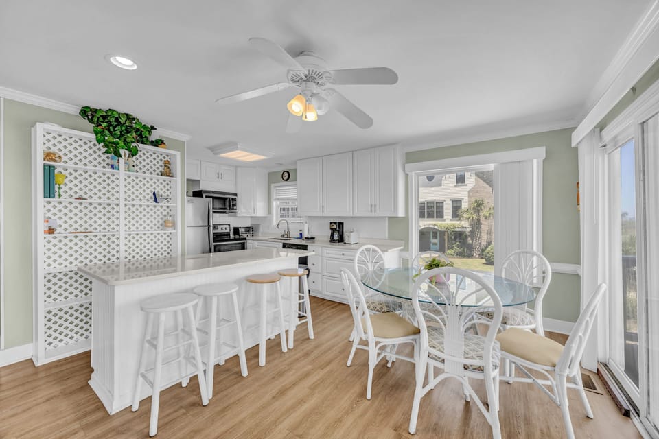 Gather around the breakfast bar or glass-top table in your bright, fully-equipped kitchen-dining area—complete with modern appliances, ceiling fan, and large windows offering natural light and serene neighborhood views.