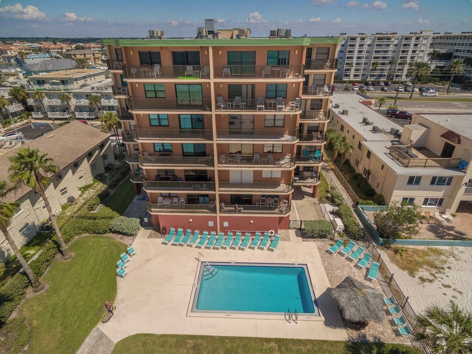 Aerial view of the beachfront property featuring a sparkling pool surrounded by turquoise lounge chairs and lush landscaping in a coastal neighborhood.