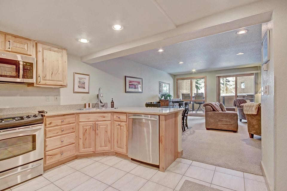 Open kitchen and living area with wooden cabinets, stainless steel appliances, and a dining table. Large windows provide natural light. Light-colored tile floors in the kitchen transition to carpet in the living area.