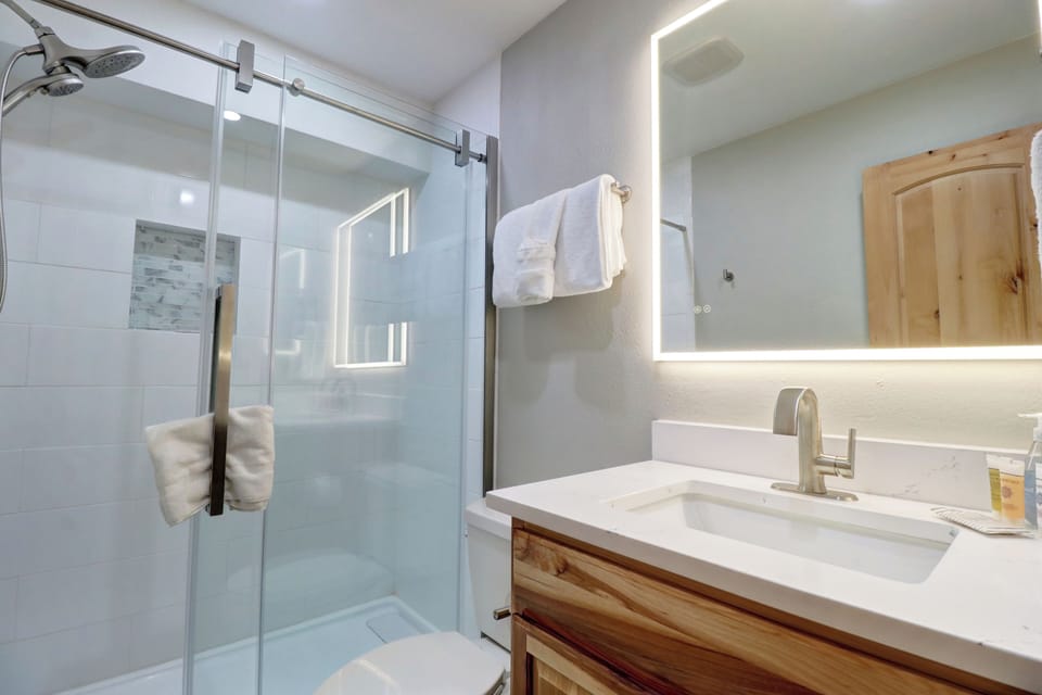 A modern bathroom features a glass-enclosed shower, a backlit mirror, a wooden cabinet, and a white countertop with a mounted faucet. Towels are hung beside the shower and on the wall.