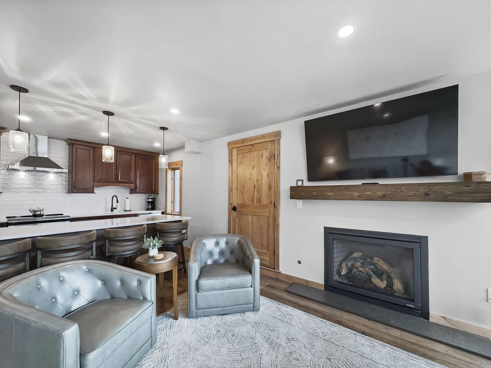 A modern living room with two gray armchairs, a wooden coffee table, a wall-mounted TV above a fireplace, and a view into the kitchen with dark cabinets and pendant lights over a countertop.
