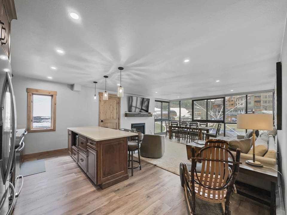 Spacious modern living area with a large kitchen island, wooden furniture, a cozy seating arrangement, and expansive windows offering a view of an outside deck and trees.