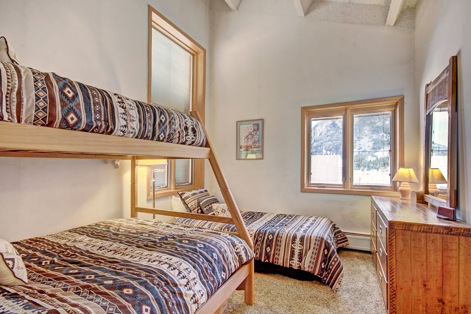 A small bedroom with a bunk bed and a single bed, both with patterned bedding. There is a wooden dresser with a mirror and two lamps. The room has two windows with mountain views.