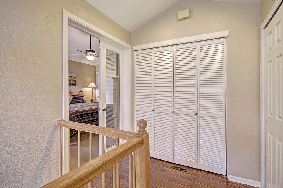 Hallway with wooden railing, bi-fold closet doors, and a view into a bedroom with a ceiling fan and bedside lamp. Walls are painted beige, and the floor has a wooden finish.