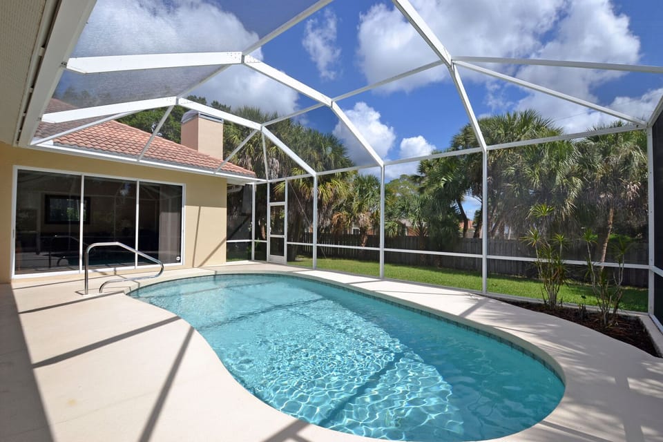 Pool and screened enclosure viewed from the pool deck, surrounded by tropical setting