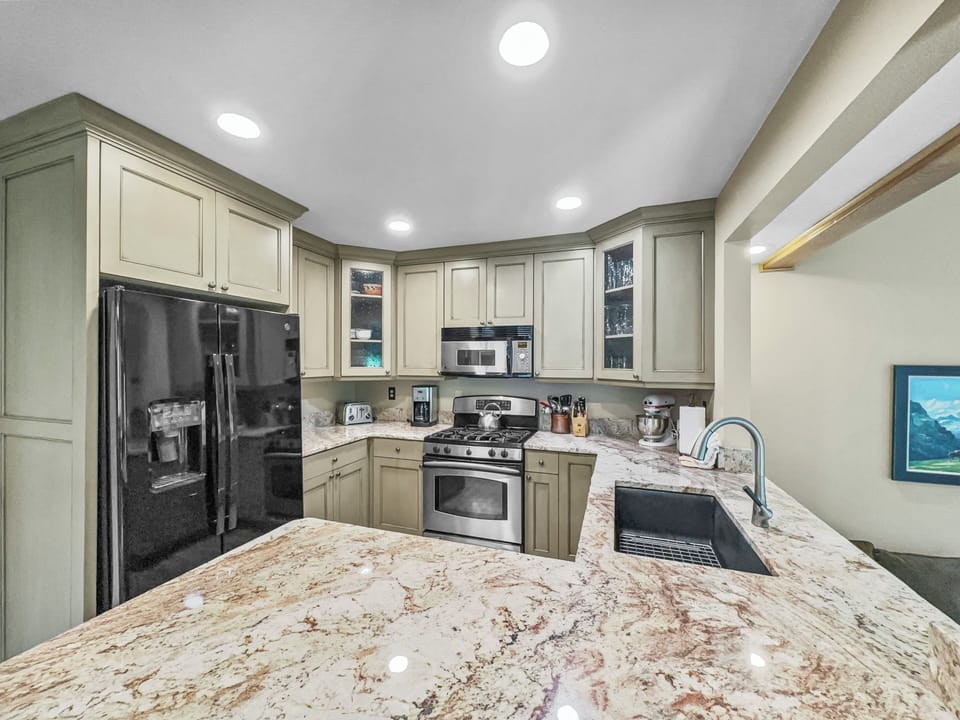 A modern kitchen with green cabinets, stainless steel appliances, a black refrigerator, a double sink, and a granite countertop. A framed picture is on the wall to the right.