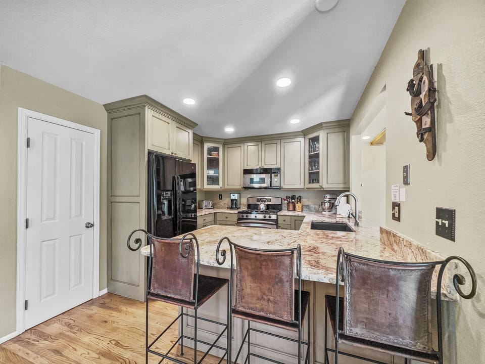 A modern kitchen with green cabinets, a granite countertop, stainless steel appliances, a black refrigerator, and three high-back leather bar stools. There is a door on the left and wood flooring.