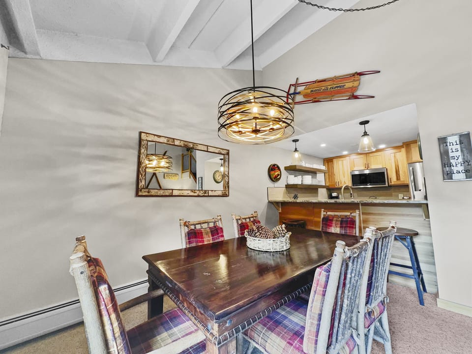 A dining area with a wooden table, plaid chairs, a large decorative mirror, and an overhead light fixture. The room adjoins a kitchen with wooden cabinets, a stainless steel fridge, and a microwave.
