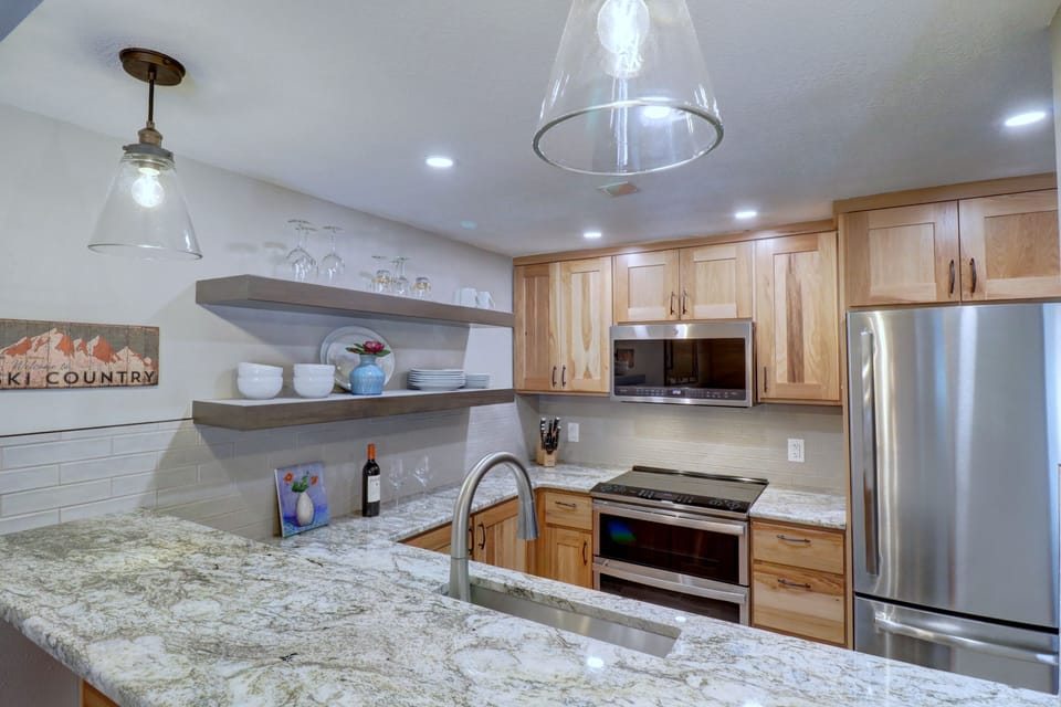 Modern kitchen with granite countertops, stainless steel appliances, wooden cabinets, open shelving with dishes, and pendant lighting. A "Ski Country" sign hangs on the wall.
