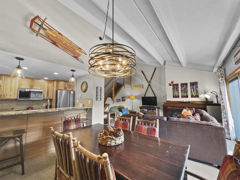 A cozy living and dining area with wooden furniture. The modern kitchen is in the background. Decor includes a chandelier, a sled hanging on the wall, and a fireplace with skis mounted above it.
