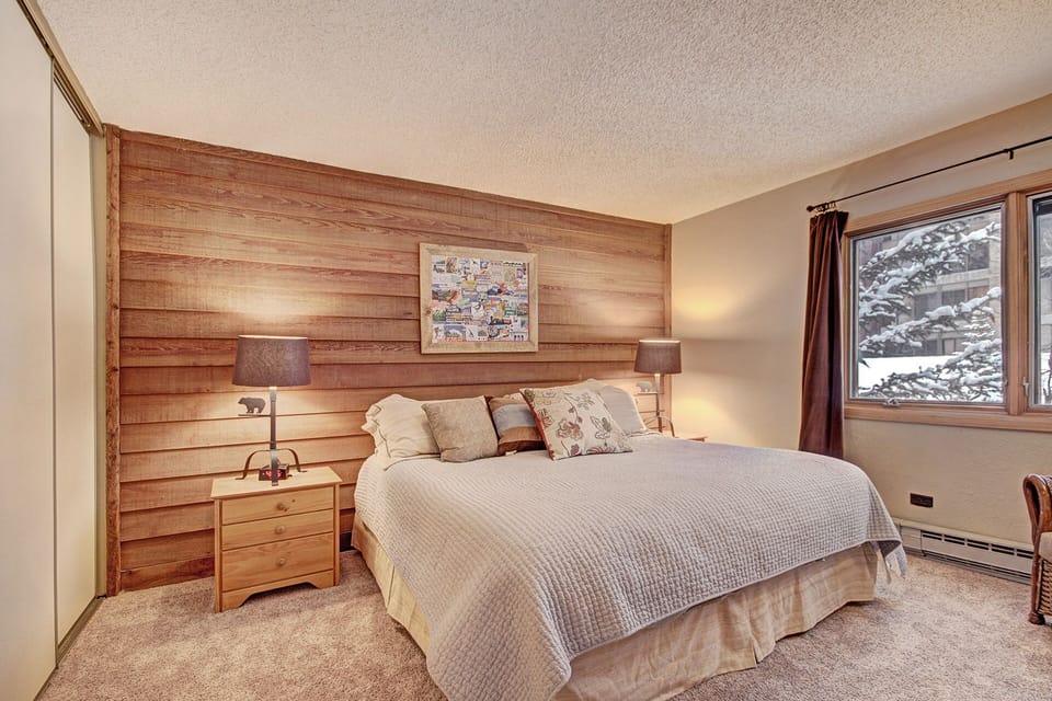 A cozy bedroom with a large bed, wooden panel accent wall, two bedside tables with lamps, framed artwork above the bed, a window with brown curtains, and a snowy view outside.