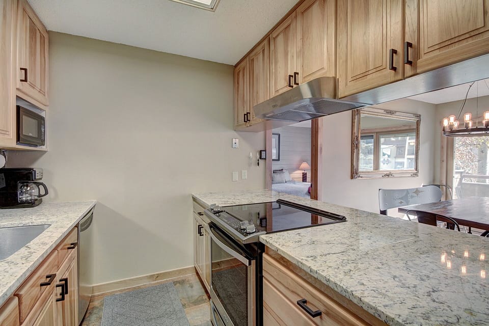 A modern kitchen with granite countertops, stainless steel appliances including a stove and microwave, light wood cabinets, and a view into a dining area with a chandelier.