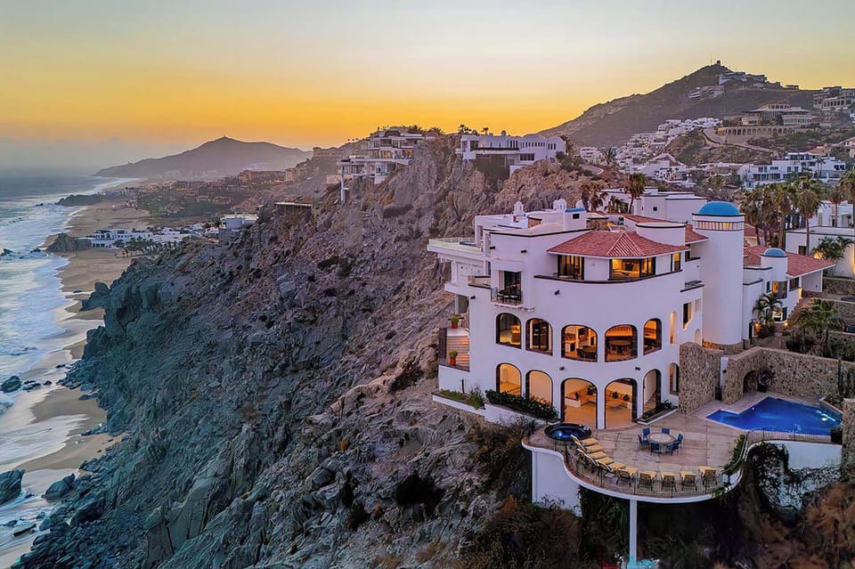 A stunning coastal estate perched on dramatic cliffs overlooking the ocean, captured during a breathtaking sunset. The property features hillside architecture, a private pool, and panoramic views of the beach and surrounding mountains.
