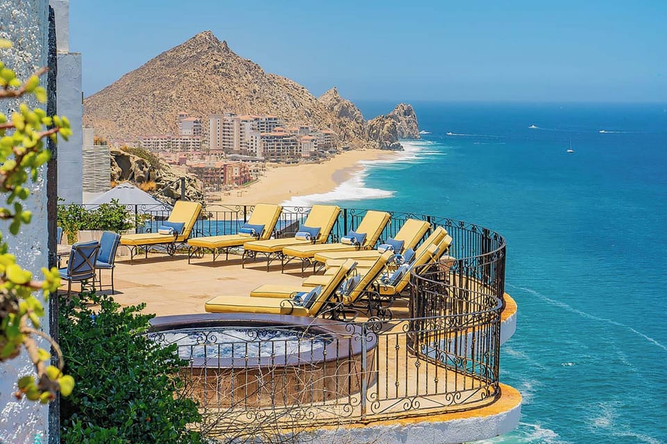 Elevated terrace featuring sun loungers and coastal panorama with dramatic rocky headland, pristine beach, and turquoise waters dotted with boats in the distance.