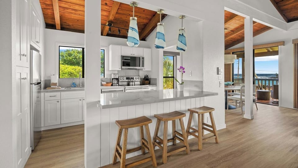Your island breakfast bar flows seamlessly into an open living space, with ocean breezes and water views visible through the open doorway to the terrace.