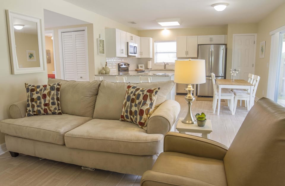 Beige sofa and armchair face the kitchen with white cabinets, stainless steel appliances, and a wooden dining table.