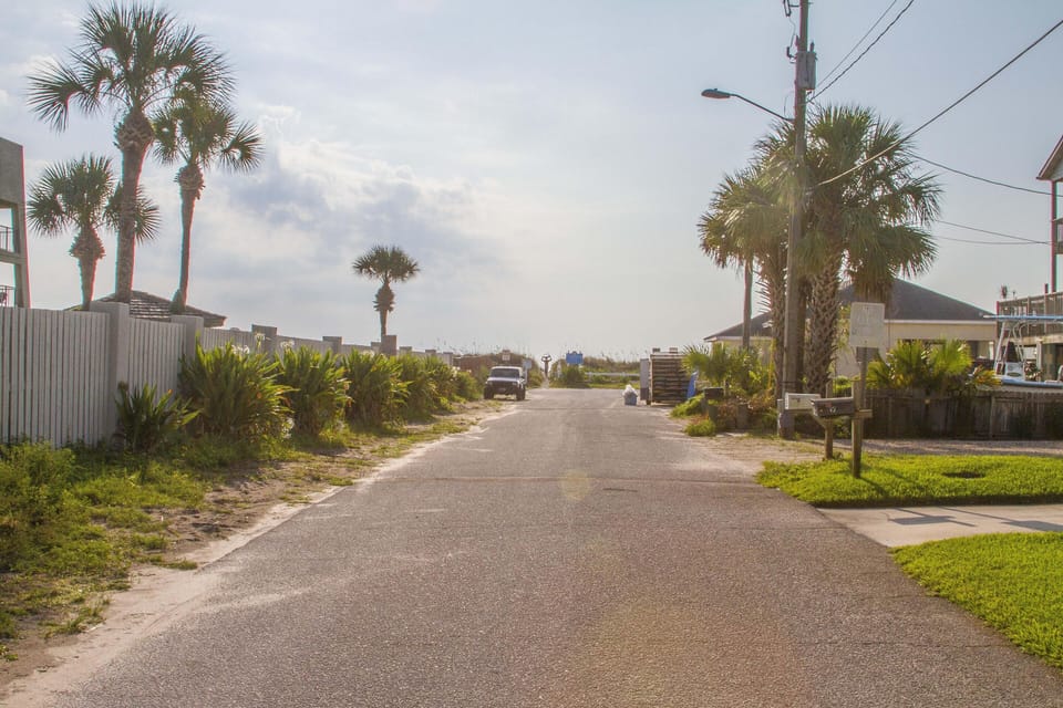 Paved street lined with palm trees and greenery leads to a sandy beach access under a partly cloudy sky.