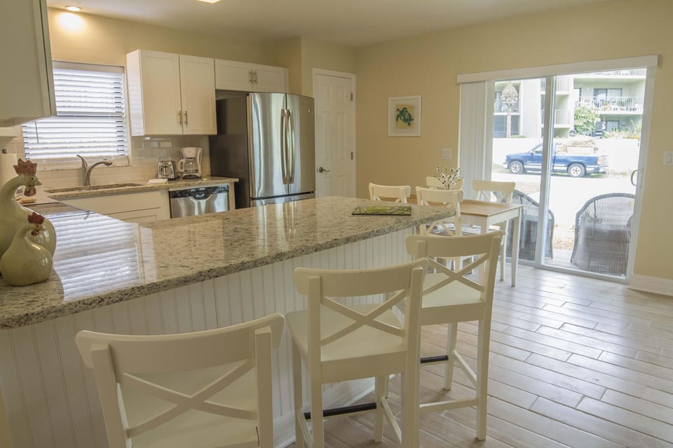 Granite-topped kitchen island with white cross-back stools faces stainless steel appliances and sliding glass door.