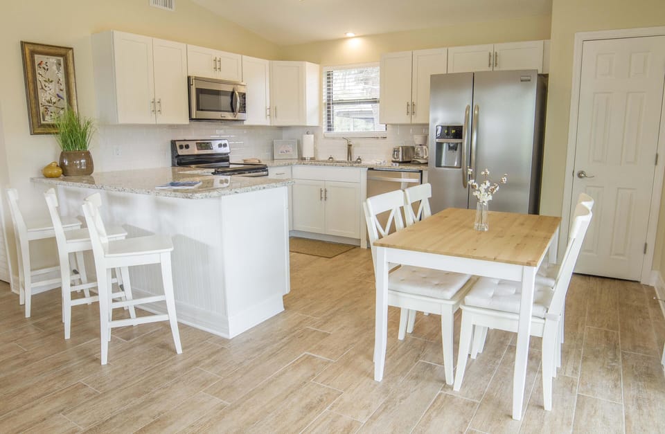 Dining area with white cushioned chairs and vase on natural wood table beside white kitchen island and stainless appliances.