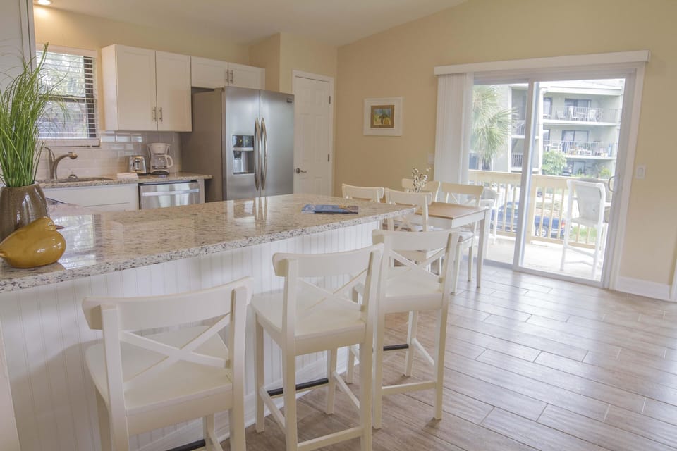 Granite countertop breakfast bar with white cross-back stools overlooks dining table and sliding balcony doors.