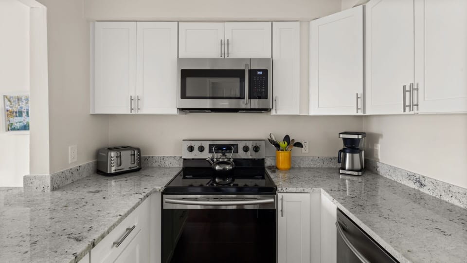 Modern kitchen with white cabinets, stainless steel appliances including a microwave, stove, toaster, and coffee maker. A yellow utensil holder and a kettle are on the counter.