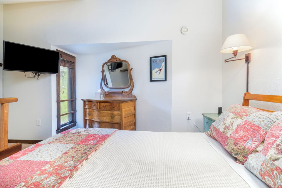 Your cozy bedroom awaits with a plush bed, charming vintage dresser, flat-screen TV, and bedside lighting. Natural light streams through the window, creating a warm and inviting retreat for your stay.