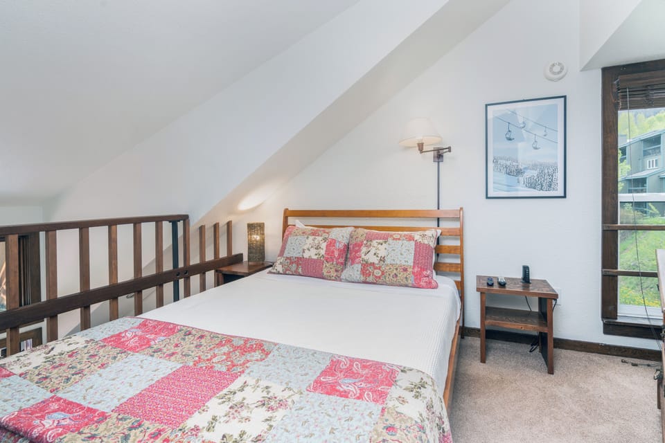 Your cozy loft bedroom awaits with sloped ceilings and mountain views, featuring a comfortable bed dressed in charming quilt bedding and a peaceful winter ski scene on the wall.
