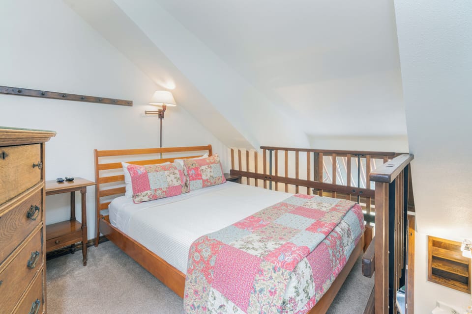 Your charming loft bedroom awaits with a cozy bed dressed in floral linens, warm lighting, and rustic wood furnishings. Perfect for a restful night's sleep.