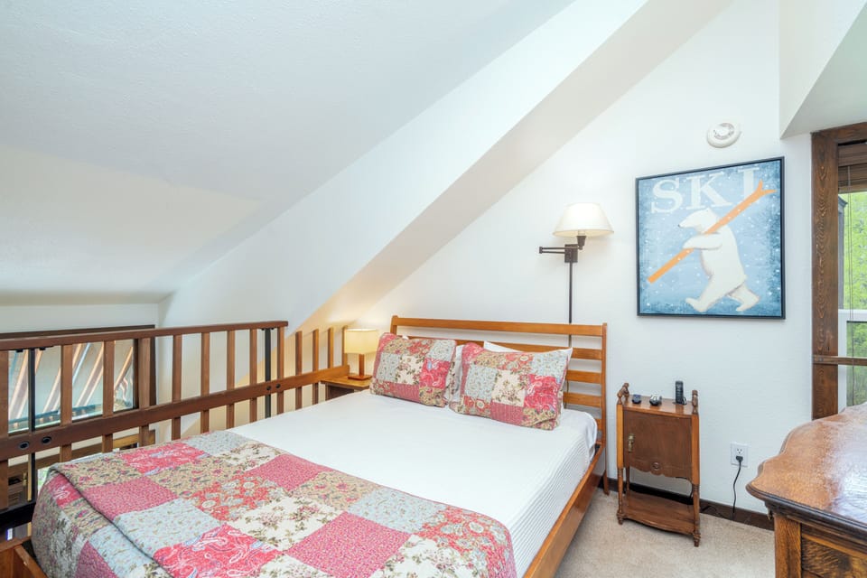 Cozy loft bedroom with vintage ski charm invites restful nights. Enjoy vaulted ceilings, warm wood accents, and a plush quilted bed awaiting your arrival.