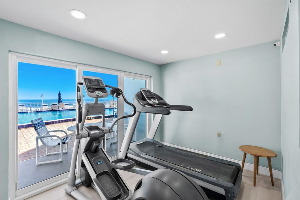 Compact fitness room featuring cardio equipment positioned beside sliding glass doors, with direct views of the pool deck and ocean beyond.