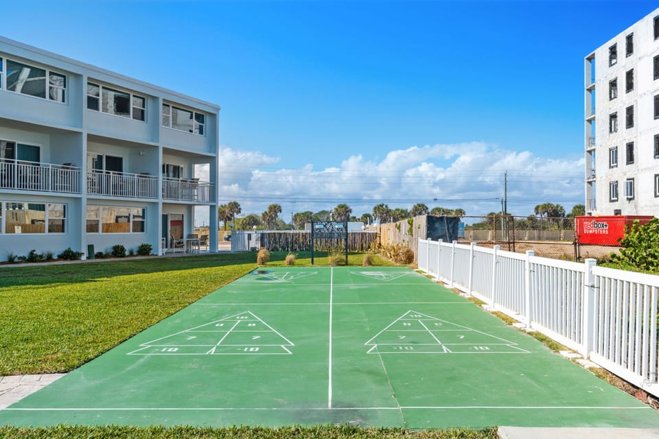 Outdoor shuffleboard court with green playing surface beside a resort building and white fence
