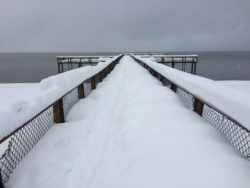 Snow-covered boardwalk extends over dark winter waters, creating a striking seasonal contrast in the surrounding area.